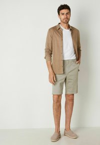 Ljusbrun knappad skjorta över en vit t-shirt, kombinerad med ljusgrå shorts och beige espadriller; rak front och avslappnad passform.