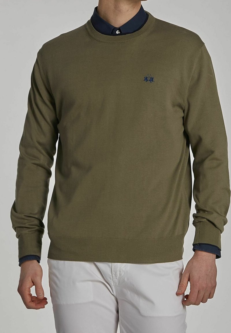 Homme portant un pull col rond vert olive par-dessus une chemise bleu marine avec un pantalon blanc, debout dans une posture décontractée devant un fond uni.