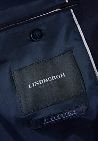 Tessuto navy con una superficie liscia presenta un bottone scuro e due etichette: "LINDBERGH BLACK" e "BISTRETCH", cucite in bianco.