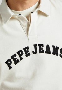 Weißes Hemd mit Kragen, auf dem "PEPE JEANS" in großen schwarzen Buchstaben über die Brust gedruckt ist, zeigt eine Nahaufnahme des Oberkörpers.