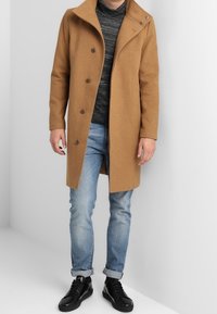 Cappotto di lana marrone con colletto a punta, sei bottoni, tasche laterali e vestibilità sartoriale, abbinato a un dolcevita grigio e jeans blu.