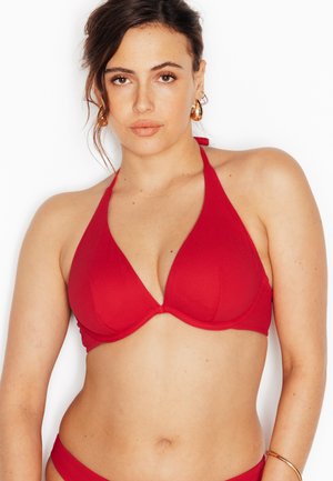 Donna che indossa un top bikini rosso a collo sciallato, con orecchini a cerchio dorati e un braccialetto su uno sfondo bianco semplice.