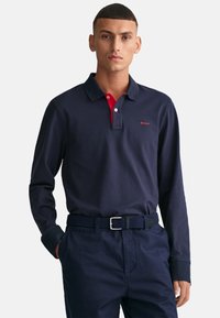 Sötétkék, hosszú ujjú póló piros gallérral, két gombbal és hímzett logóval. Sötét nadrággal és texturált szövet övvel viselve.