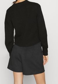 Czarny dziany sweter z okrągłym dekoltem i prążkowanymi mankietami, zestawiony z czarnymi szortami z zamszu. Tkanina ma fakturowany wygląd.