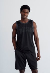 Svart atletisk tanktopp med hevet "ELLESSE"-logo og kontrastfarger, kombinert med svarte shorts. Glatt tekstur, tettsittende design.