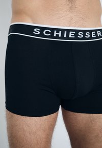 Crne pamučne bokserice s bijelim elastičnim pojasom na kojem se nalazi logo "Schiesser". Glatka tekstura i prilagođeni dizajn.