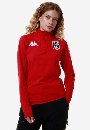 Maglietta sportiva rossa a maniche lunghe con colletto a mezza zip, con logo Kappa bianco ed emblema dello sci statunitense sul petto. Texture liscia.