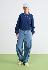 Tummansininen huppari, jossa on punainen sydänlogo, ylisuuria vaaleansinisiä cargo-housuja ja kermaisia lenkkareita, käytettynä vaaleansinisen lippiksen kanssa.