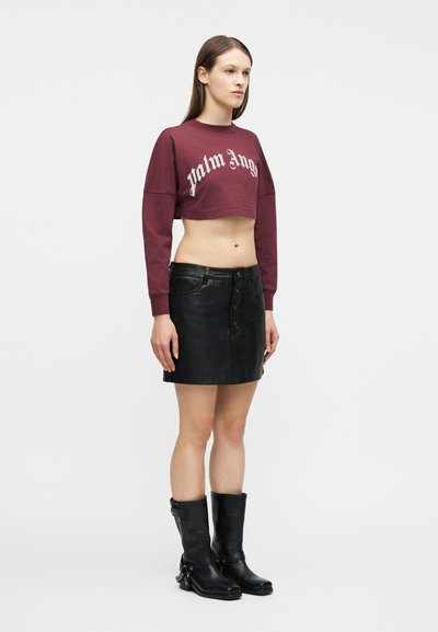 Γυναίκα που φοράει κοντό μπουφάν μπορντό, μαύρη δερμάτινη μίνι φούστα με κουμπιά μπροστά και μαύρες μπότες μέχρι τη μέση της γάμπας, στέκεται μπροστά σε λευκό φόντο.