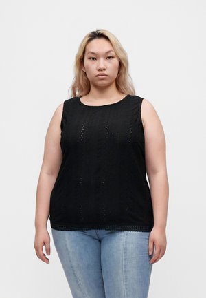 Jonge vrouw met blond haar die een zwarte mouwloze top en lichtblauwe spijkerbroek draagt, staand tegen een effen lichte achtergrond.