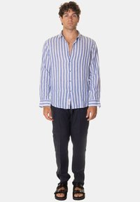 Camicia di lino leggera a righe verticali blu e bianche, con chiusura a bottoni sul davanti e maniche lunghe, abbinata a pantaloni scuri e sandali.