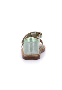 Sanda apare metálica verde, vista trasera. Cuenta con una parte superior suave, una correa ajustable y una suela plana marrón. Textura suave y diseño sencillo.