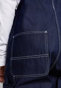 Laivastonsiniset farkkuhaalari, joissa valkoiset saumat, suuri sivutasku vasemmalla ja sileä pinta.