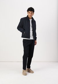 Marineblauwe jas met een rits, staande kraag en subtiele rode accenten; gedragen met zwarte broeken en beige schoenen op een neutrale achtergrond.