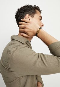 Pulsera trenzada negra con eslabones de metal en tono dorado. Se lleva en la muñeca, complementada con una camisa beige claro abotonada. Primer plano de los detalles.