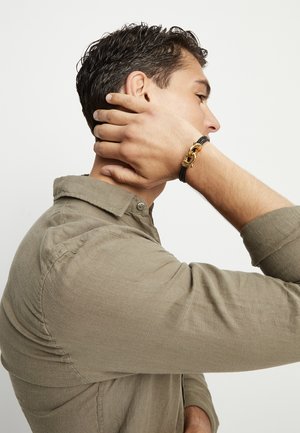 Pulsera trenzada negra con eslabones de metal en tono dorado. Se lleva en la muñeca, complementada con una camisa beige claro abotonada. Primer plano de los detalles.