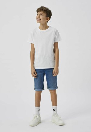 T-shirt en coton blanc, short en denim bleu avec des revers, baskets blanches et chaussettes blanches avec un logo noir. Tenue simple et décontractée.