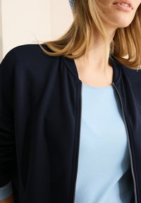 Giacca zip-up blu navy con scollo rotondo, indossata sopra una maglietta leggera azzurro chiaro a maniche corte. Entrambi i capi presentano un tessuto morbido e liscio.