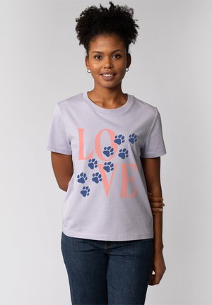 Frau mit lockigem Haar lächelt, trägt ein lavendelfarbenes T-Shirt mit korallenrotem "LOVE"-Schriftzug und blauen Pfotenabdrücken, kombiniert mit blauen Jeans.