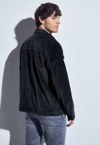 Černá kordurová bunda s uvolněným střihem, která má zádový yoke a dvě boční kapsy, v kombinaci s světle šedými džíny.