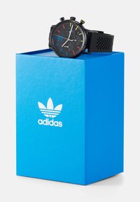 Reloj cronógrafo negro con acentos coloridos en una correa negra texturizada, descansando sobre una caja azul brillante con un logo blanco de Adidas.