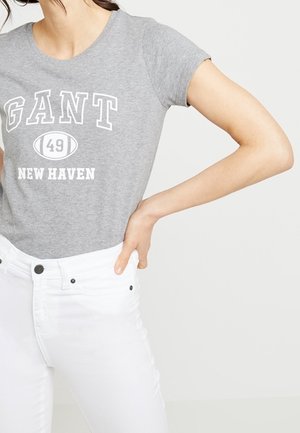 Woman wearing a grey GANT T-shirt with "49 New Haven" text and white high-waisted jeans, hand resting on hip.