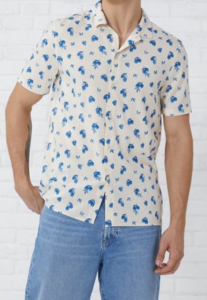 Homme portant une chemise beige à manches courtes avec un motif de feuilles bleues, associée à un jean bleu clair, debout contre un mur en briques blanches.