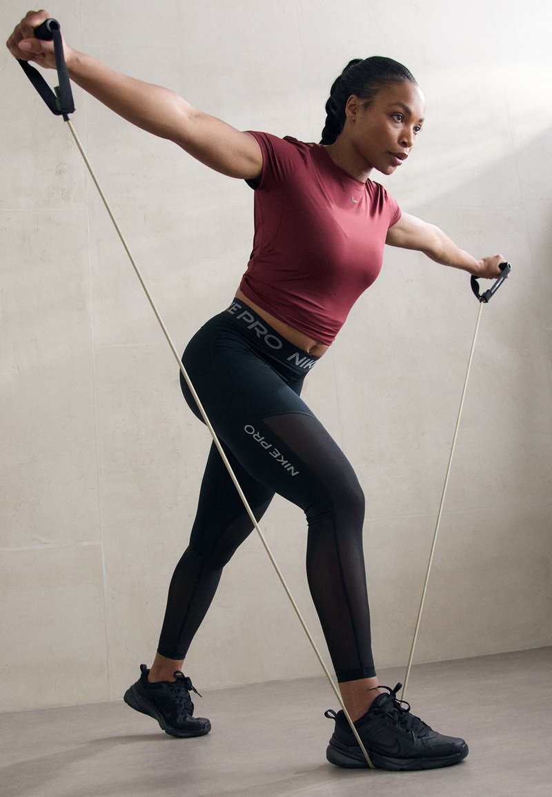 T-shirt de sport rouge et leggings noirs avec des accents en maille. En train de faire de l'exercice avec une bande de résistance dans un environnement aux tons neutres. Baskets noires.