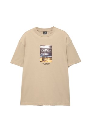T-shirt en coton beige à manches courtes, arborant un graphique central d'une scène désertique avec un cactus et une route, accompagné de texte en dessous.