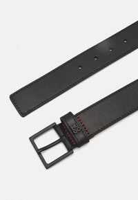 Ceinture en cuir noir avec une boucle noire mate, des accents de couture rouge et plusieurs trous pour un ajustement de taille.