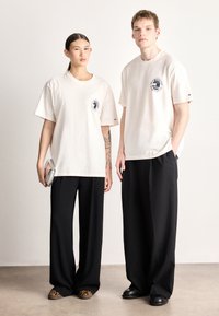 Svetlo bež ohlapne bombažne majice z logotipskimi našivkami, kombinirane s širokimi črnimi hlačami. Ena oseba drži srebrno torbico, druga pa ima viden tatu.