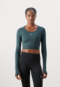 Crop top lung cu maneci lungi de culoare verde închis, cu decolteu rotund și logo-ul alb Nike; asortat cu colanți negri cu talie înaltă. Textura netedă.