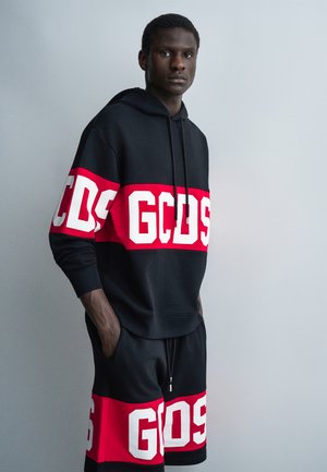 Άνδρας με τα χέρια στις τσέπες φοράει μαύρο φούτερ με κουκούλα και σορτς με έντονα κόκκινα και λευκά λογότυπα GCDS κατά μήκος του στήθους, των χεριών και των ποδιών.