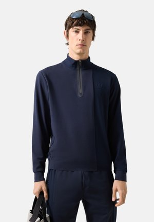 Άνδρας που φοράει ναυτικό μπλε πουλόβερ με φερμουάρ και παντελόνι που ταιριάζει, κρατώντας μαύρη και λευκή τσάντα, με γυαλιά ηλίου που ξεκουράζονται πάνω σε κοντά καστανά μαλλιά.