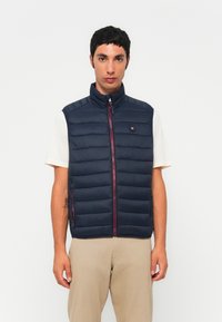 Marineblå puffer vest med quiltedesign, høy krage og rød glidelås. Lett materiale med sidelommer og logo-patch på brystet.
