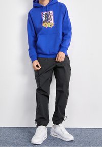 Modrá mikina s grafikou a přední kapsou, spárovaná s černými cargo kalhotami a bílými teniskami s vzduchovým polstrováním.