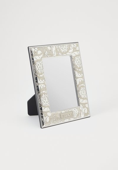Lustro Versace z wzorzystą srebrną ramą, zdobione misternymi kwiatowymi wzorami, o prostokątnym kształcie, z czarnym stojakiem dla podparcia.