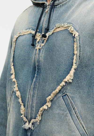 Τζιν μπουφάν με σκισμένο άνοιγμα σε σχήμα καρδιάς, μπλε ύφασμα, κουκούλα και σκούρα κορδόνια. Διαθέτει ξεφτισμένα άκρα και πλαϊνές τσέπες.