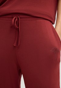 Bordeauxfarbene Jogginghosen aus einer weicheren Baumwollmischung mit einstellbarem Kordelzug. Mit einem kleinen, mit Strass verzierten Blumenmotif in der Nähe der Tasche.