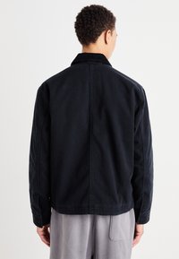 Svart denimjakke med kordfløyelskrage. Har en avslappet passform og lange ermer. Bak har den en søm som går vertikalt nedover midten.