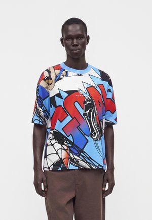 Άνδρας που φοράει πολύχρωμο γραφικό t-shirt με έντονα κόκκινα, μπλε, μαύρα και λευκά αφηρημένα αθλητικά σχέδια και καφέ παντελόνι, στέκεται μπροστά σε απλό φόντο.
