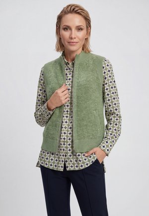 Donna che indossa un gilet verde con cerniera sopra una camicia a maniche lunghe a fantasia e pantaloni scuri, in piedi con una mano in tasca e l'altra che tiene il gilet.