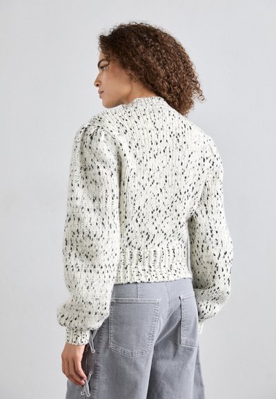Chunky Strickpullover in Weiß mit schwarzen Sprenkeln, kurze Form, gerippter Saum und Puffärmel. Getragen mit grauen Hosen.