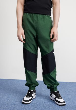 Persona che indossa pantaloni sportivi Nike verdi e neri e scarpe da ginnastica nere e bianche in piedi su un tappeto blu.