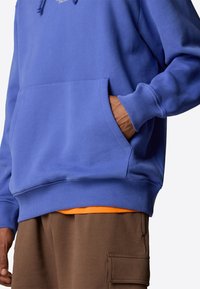 Blue hooded sweatshirt with a front pocket, ribbed cuffs and hem, worn over an orange shirt, paired with brown sweatpants featuring pocket detail.