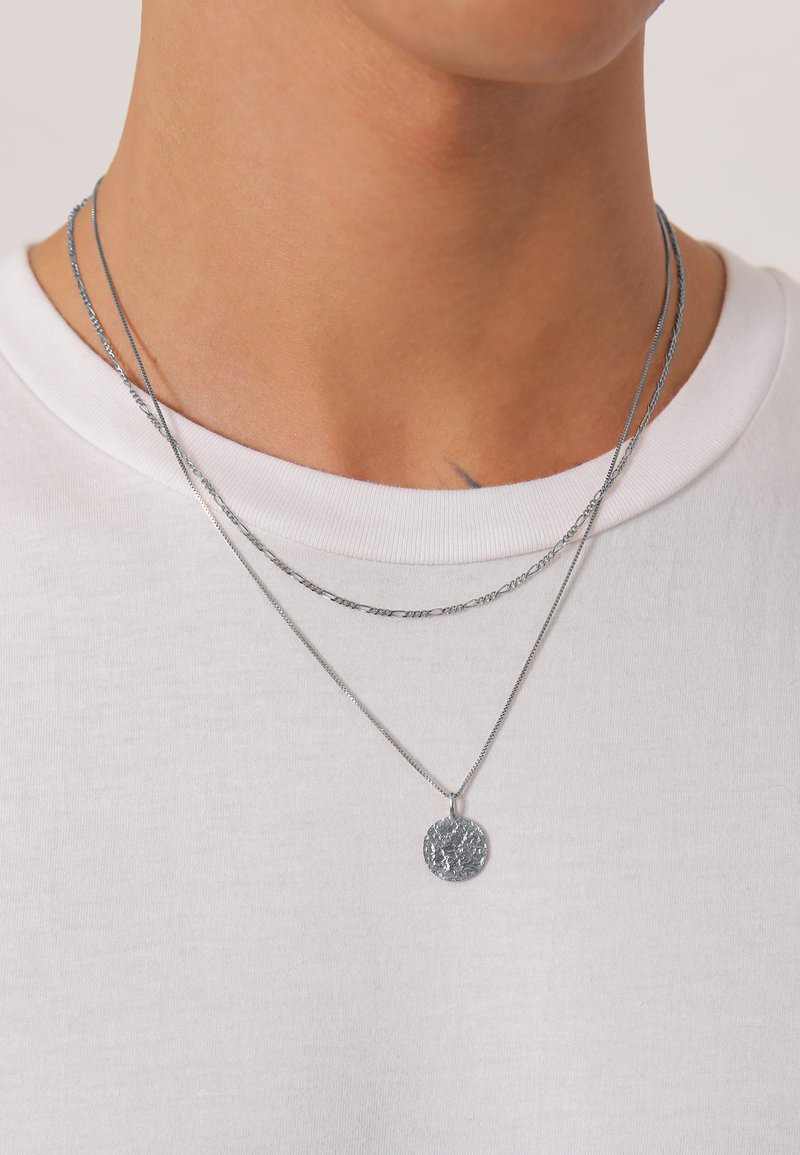 Des colliers superposés en argent avec une fine chaîne et un pendentif circulaire texturé, portés sur un t-shirt blanc à col rond.