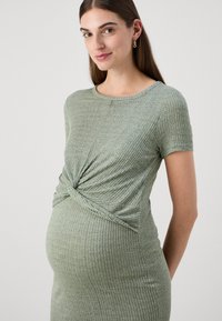 Vihreä poikittain ribattu äitiysmekko, jossa on lyhyet hihat ja solmittu yksityiskohta vyötäröllä. Pehmeä materiaali, kapeasti istuva muotoilu.