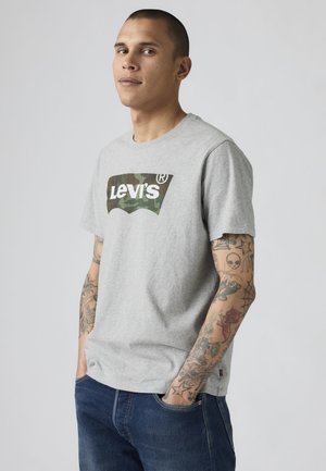 Jeune homme aux cheveux courts portant un t-shirt gris Levi's et un jean bleu, montrant ses bras tatoués, debout les mains dans les poches devant un fond uni.