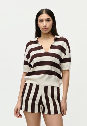 Nő laza szabású kötött felsőben, amelyen széles, vízszintes barna és fehér csíkok futnak, valamint hozzáillő kötött rövidnadrágban, amelyen függőleges csíkok találhatók.