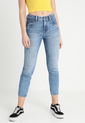 Femme portant un jean taille haute bleu clair, un débardeur court jaune et des baskets noir et blanc, debout devant un fond blanc.